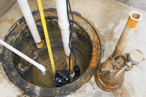 An old sump-pump has been replaced with a new one. The old pump sits outside of a sump pit, while the new one has been installed. PVC pipe extends outward as a means to expel water from the residence.