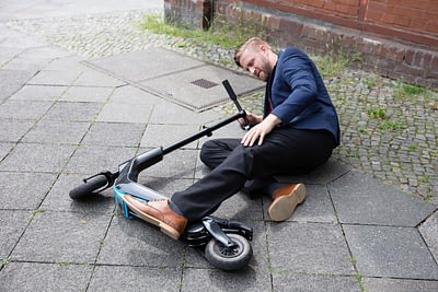 young man in business suit lying on ground with electric scooter under his left leg