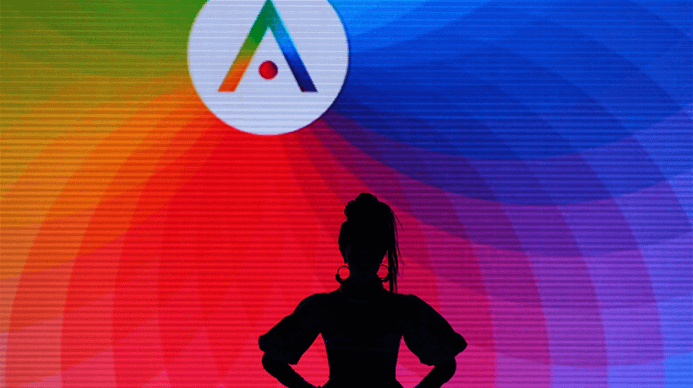 An outline of a woman with large earrings against a rainbow colored LED board with the aveda logo