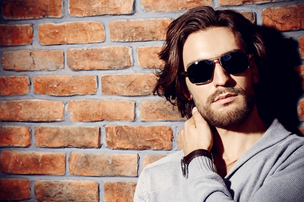 stylish man with long hair and beard wearing sunglasses in front of a brick wall