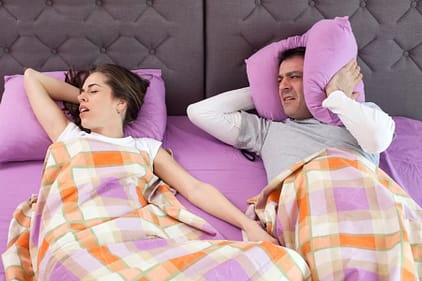 couple in bed with woman snoring and man covering his ears with pillows
