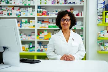 Pharmacy technician smiles behind the counter with shelves of medicine in background
