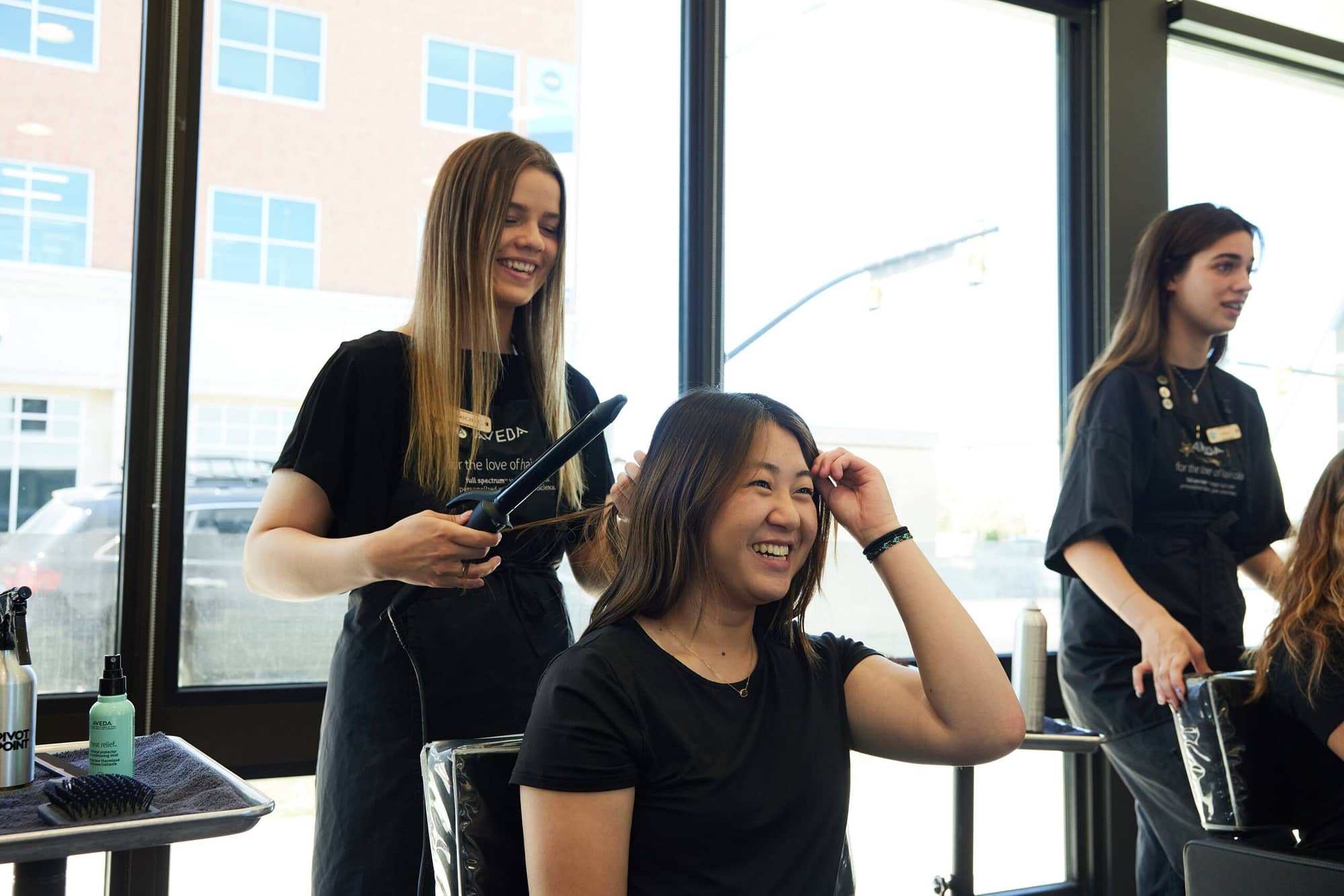 Beauty school student laughs with client sitting in chair while administering curling iron