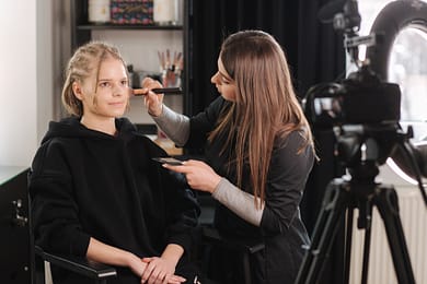 Makeup artist applying blush to woman seated in front of camera