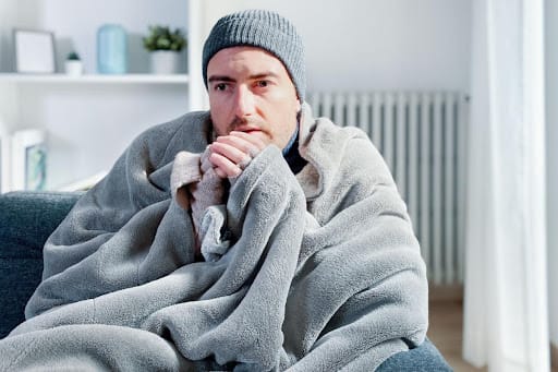 A man huddles up with a blanket and hat on his sofa because his heater isn't working. The blanket and hat are both gray colored. There are shelves in the background.