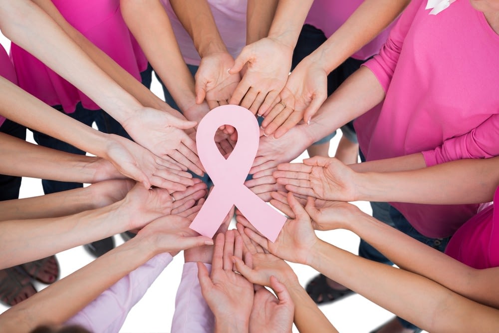 A circle of hands holding a pink breast cancer awareness ribbon.