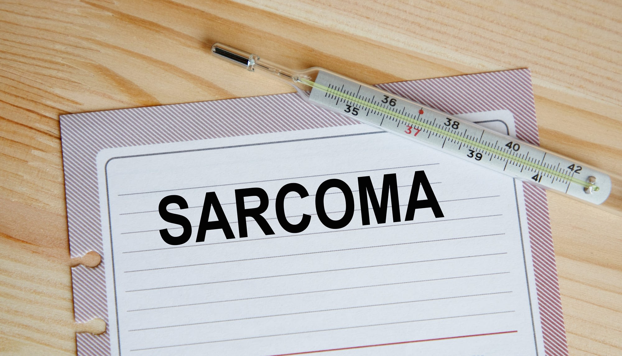 On the sheet is the inscription SARCOMA, next to the thermometer, against the background of a wooden table. A medical concept.