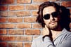 stylish man with long hair and beard wearing sunglasses in front of a brick wall
