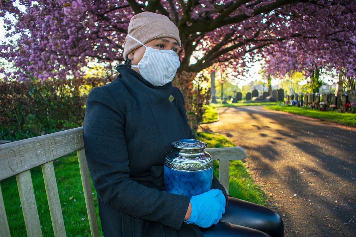 woman wearing a mask sitting on a park bench in a cemetery holding an urn