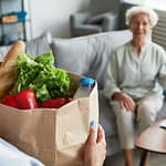 Caregiver delivering bag of groceries to smiling client at their home