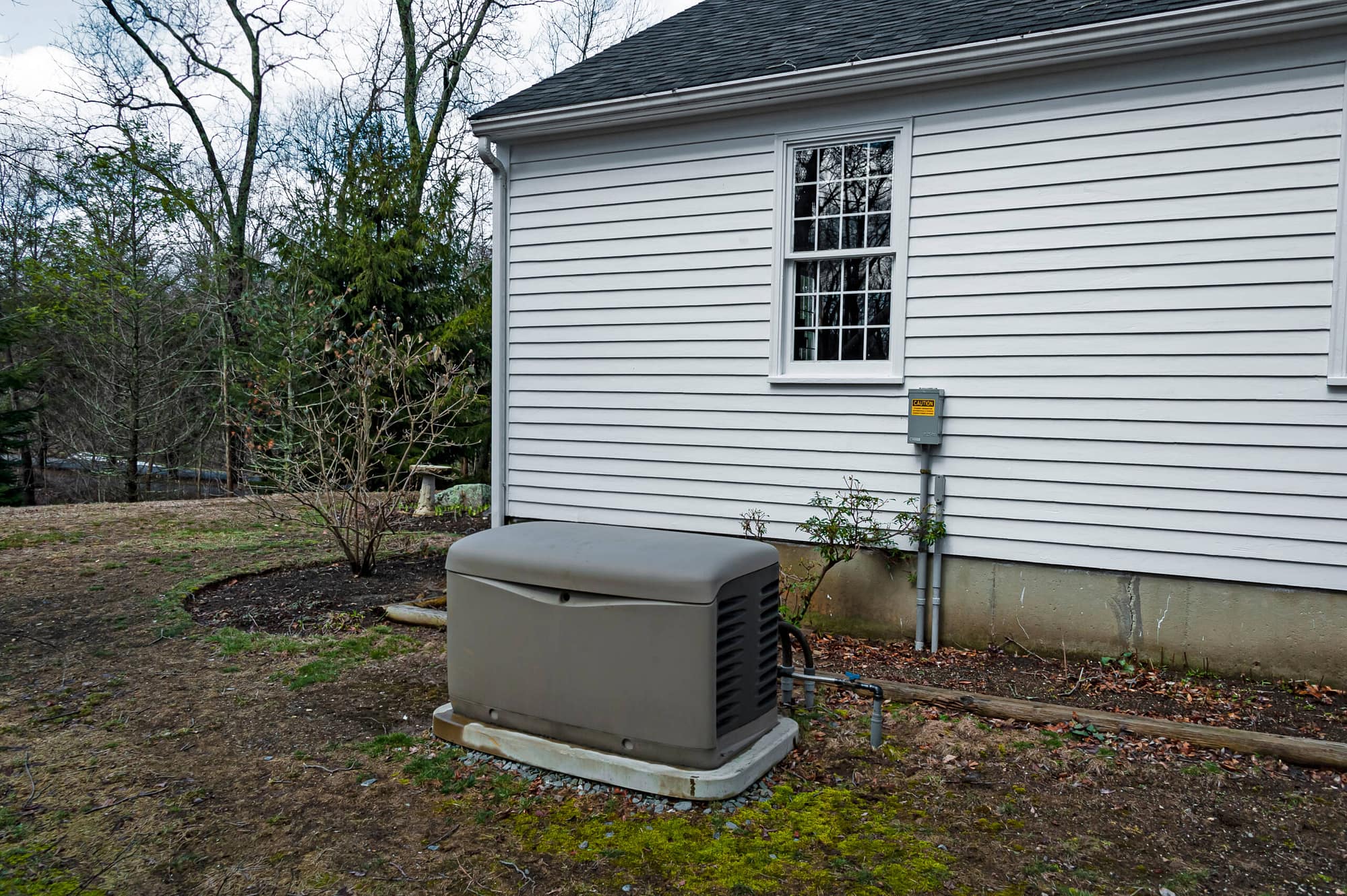 A representation of the big factors to consider when finding the cost to install a Generac generator: The generator, cement pad, wiring, and transfer switch, outside a home.