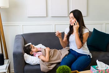 Parent on phone with doctor reads thermometer sitting by sick child on sofa