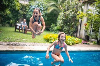 Young children jumping into backyard swimming pool as guardian watches on 