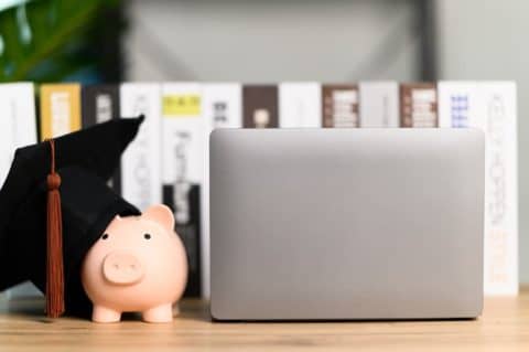 A piggy bank with a graduation cap next to a laptop computer