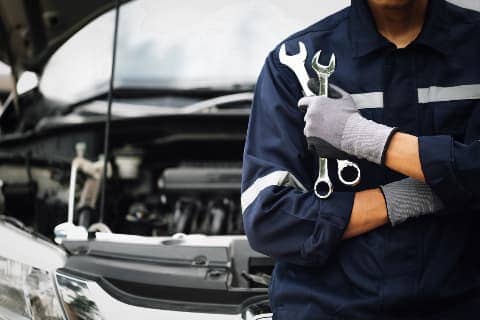 Car mechanic holding two wrenches and leaning against front bumper of car with hood up