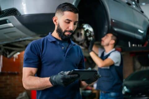 Supervisor at car shop checking tablet while mechanic works in background on car