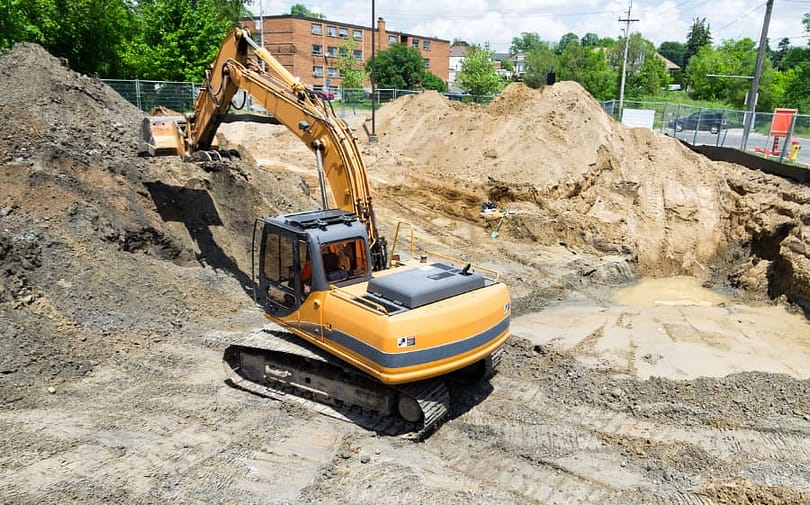 A large construction excavator removes contaminated soil from an urban brownfield development site